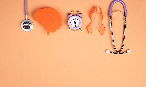 Bild zum Tag der Multiplen Sklerose. Zu sehen sind ein Stetoskop, die orangene Gedenkschleife, ein Wecker und ein ausgeschnittenes Papier-Gehirn.