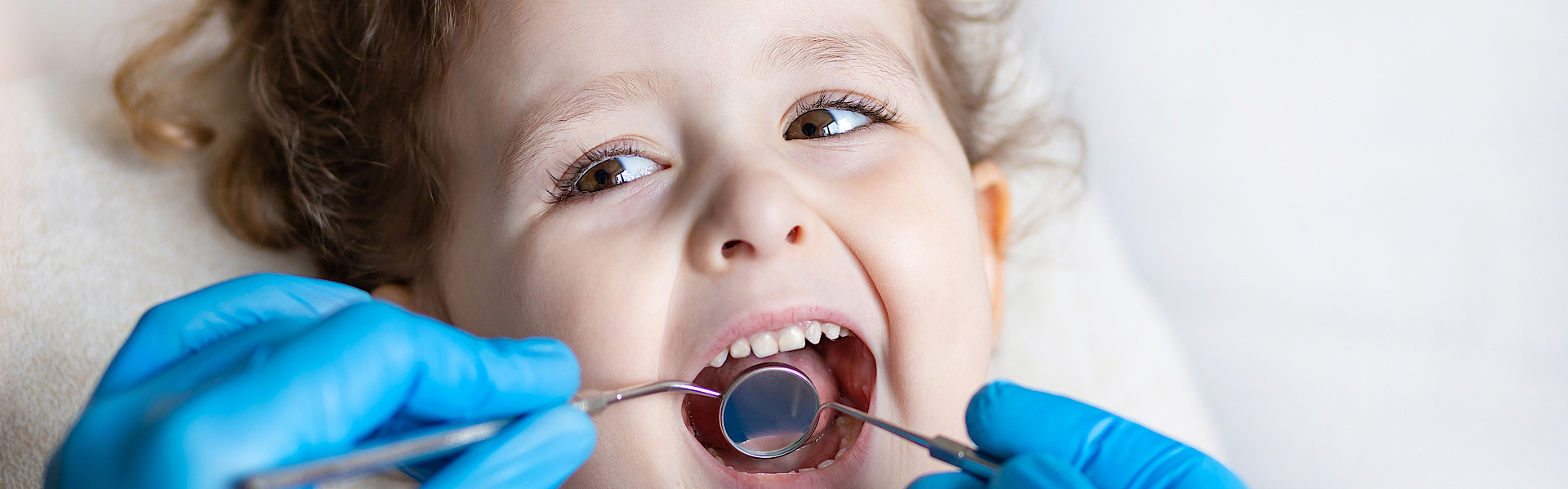 Ein kleines Mädchen mit braunen Locken liegt auf dem Untersuchungsstuhl beim Zahnarzt. Sie schaut den Arzt an. Er untersucht mit dem Spiegel ihre Milchzähne.
